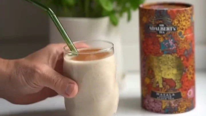 Ręka trzyma szklankę z kremowym smoothie i metalową słomką, obok kolorowa puszka herbaty Adalbert's