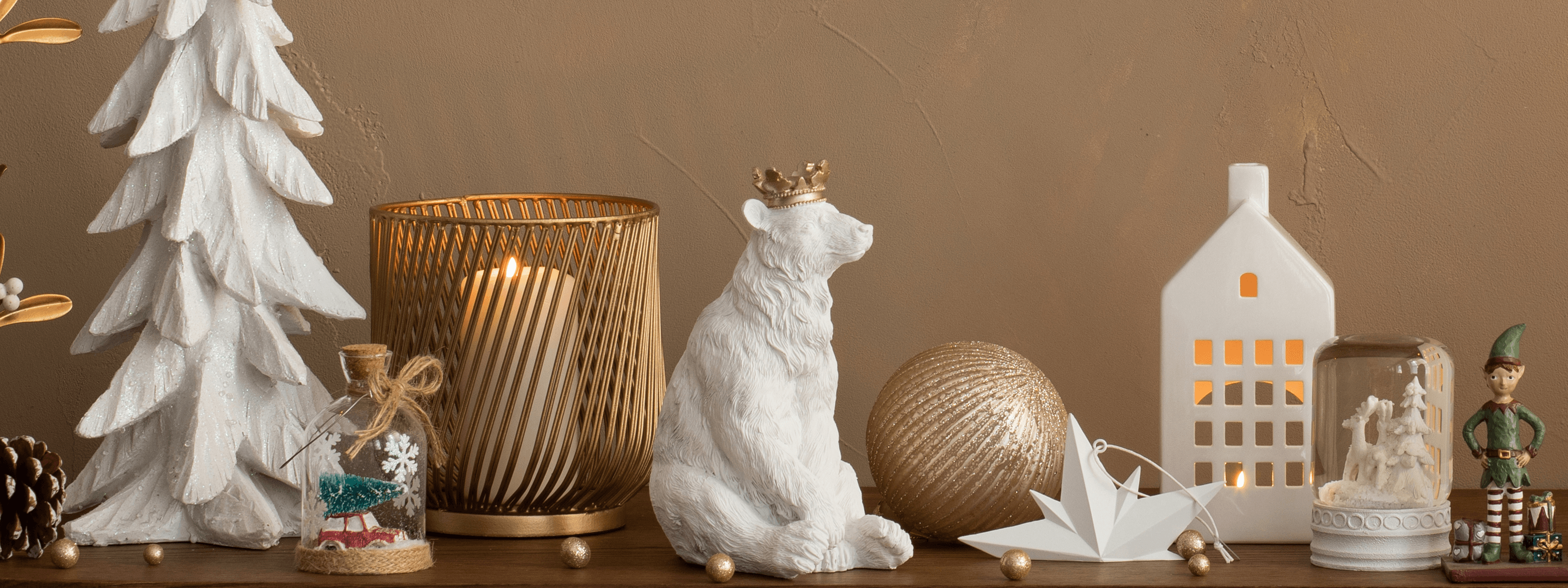 Świąteczne dekoracje na stole: biała figurka misia z koroną, złoty świecznik, dom z lampionem, choinka i elf