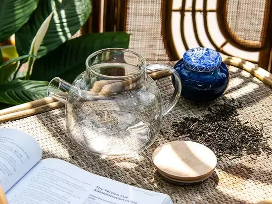 Szklany dzbanek do herbaty na lnianej tkaninie obok sypanej czarnej herbaty, ceramicznego pojemnika i otwartej książki