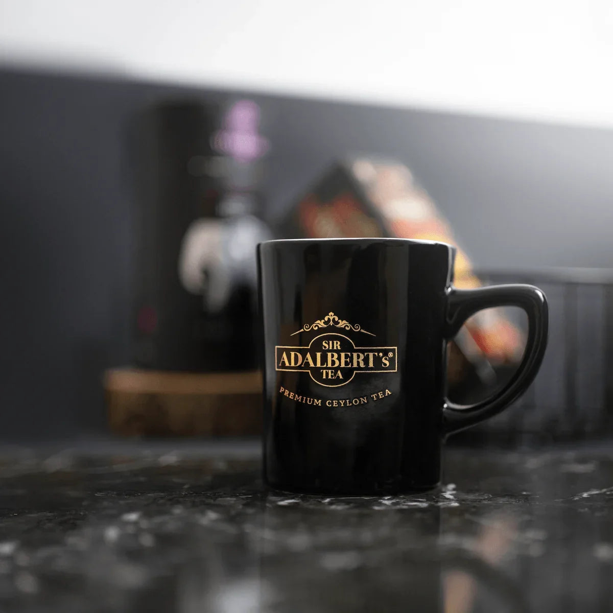 Czarny kubek z logo Sir Adalbert's Tea na marmurowym blacie, w tle rozmyte opakowania herbaty