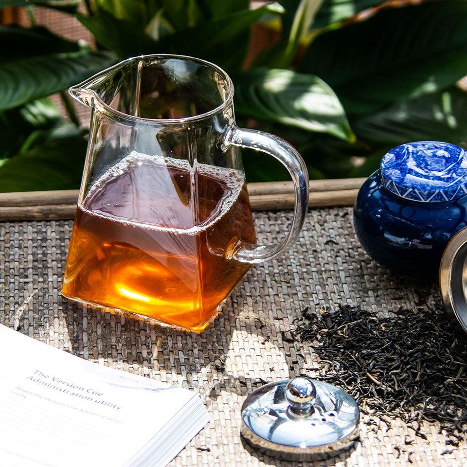 Szklany dzbanek z zaparzaną herbatą na wiklinowej powierzchni obok niebieskiego pojemnika i liści herbaty