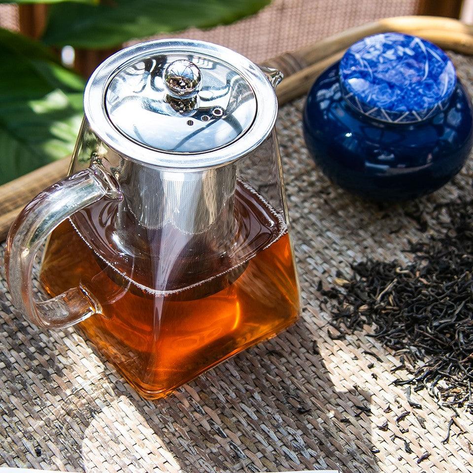 Szklany czajnik z zaparzoną herbatą, metalowy filtr, niebieski pojemnik i liście herbaty na wiklinowej tacy