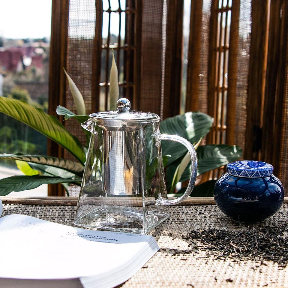 Szklany czajnik z metalowym zaparzaczem na tle roślin i bambusowych paneli, obok niebieski pojemnik i liście herbaty