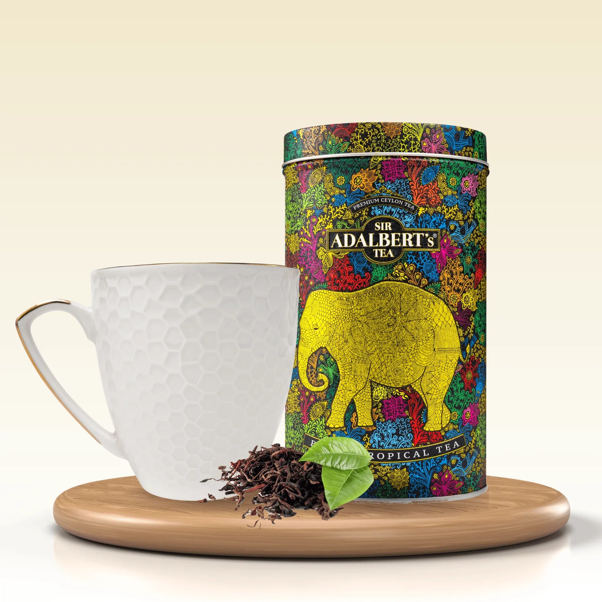 Puszka Sir Adalbert's Tropical Black Tea z kolorowym wzorem i słoniem, biały kubek, liście herbaty na drewnianej tacy