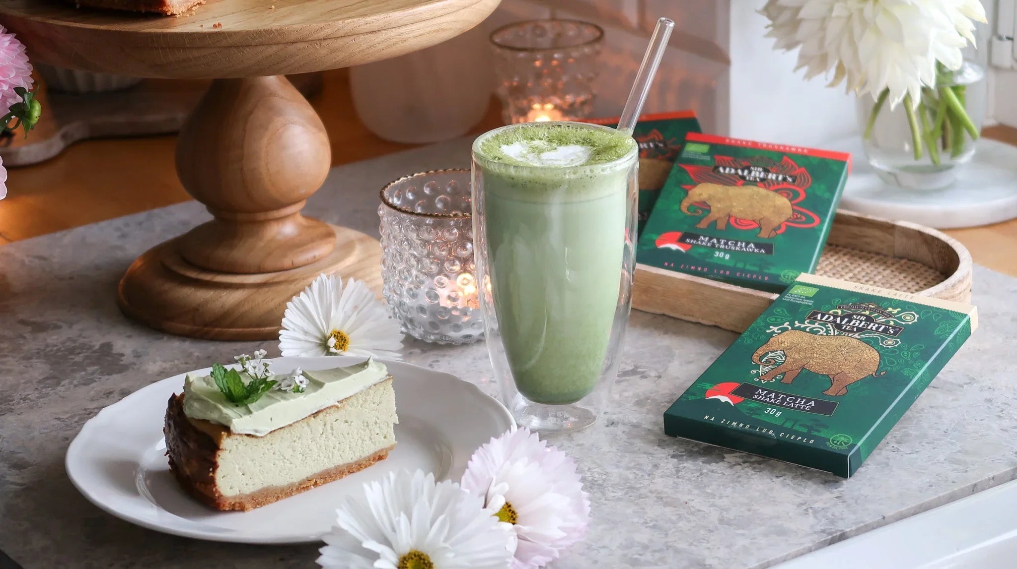 Szklanka matcha latte ze słomką, kawałek sernika na talerzu, opakowania herbaty matcha na stole