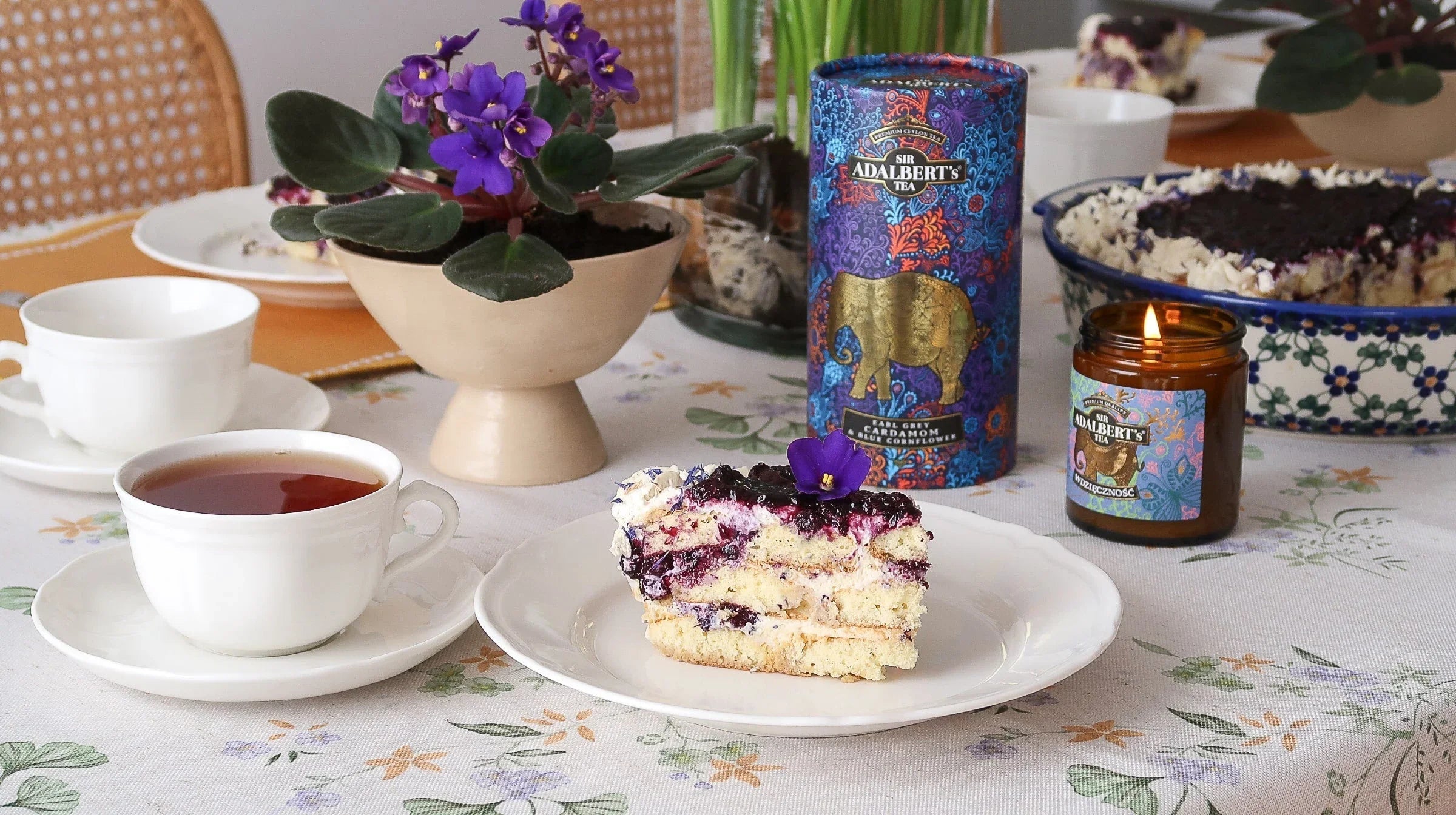 Filiżanka herbaty, deser z jagodami na talerzu, kwiaty w doniczce i opakowanie herbaty Sir Adalbert's na stole