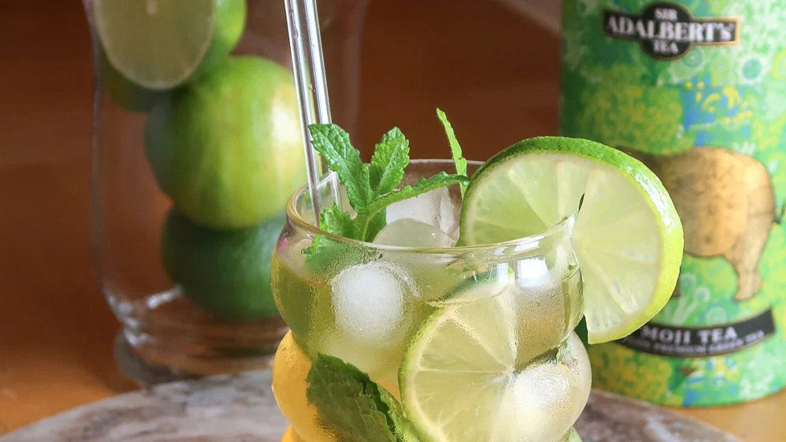 Szklanka z lodem, plasterkami limonki i miętą na tle zielonej puszki herbaty Adalbert's Mojii Tea i słoika z limonkami