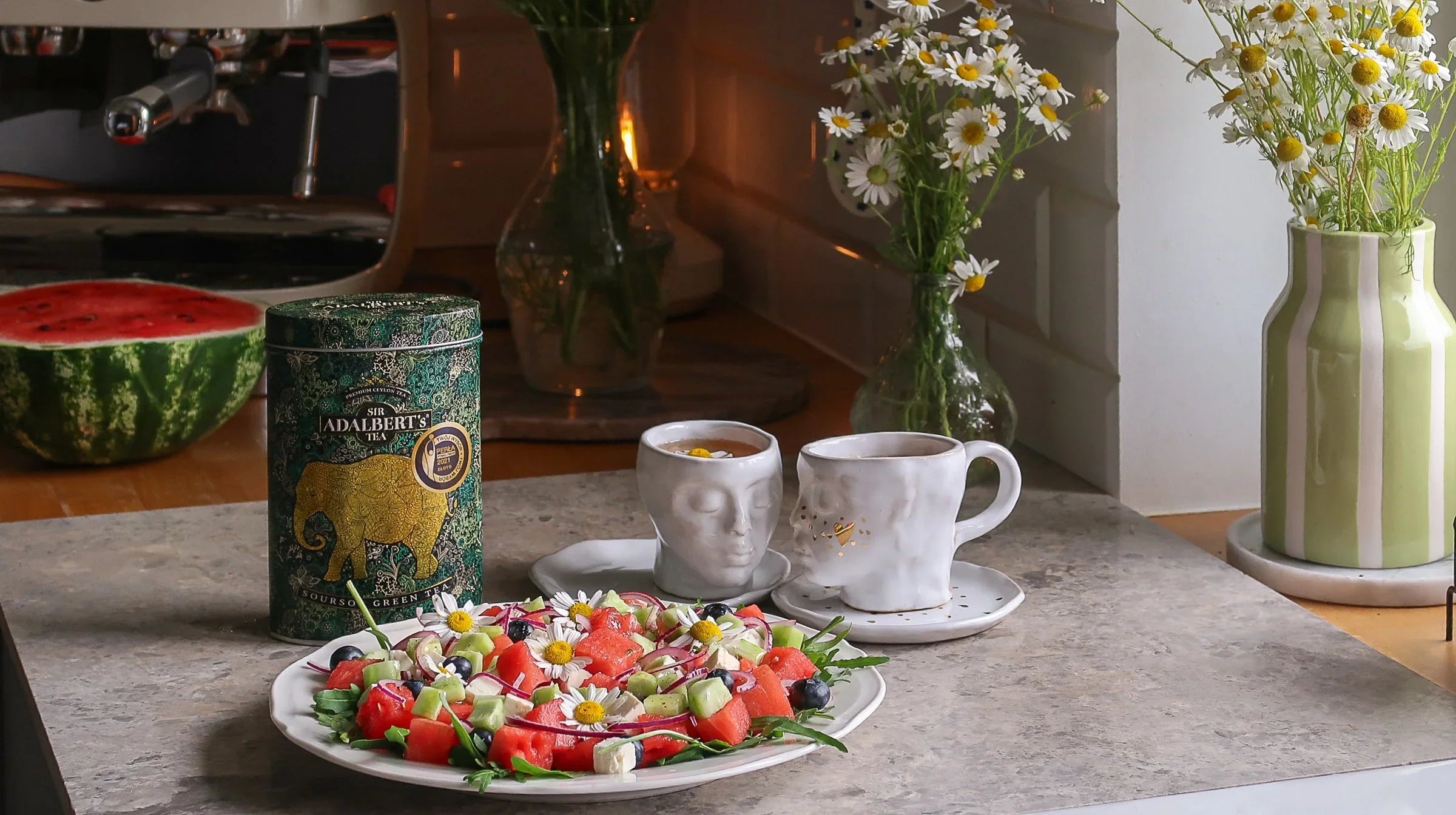 Sałatka z arbuzem, ogórkiem i kwiatami stokrotki na talerzu, herbata w kubkach, puszka zielonej herbaty, kwiaty w wazonach na blacie kuchennym