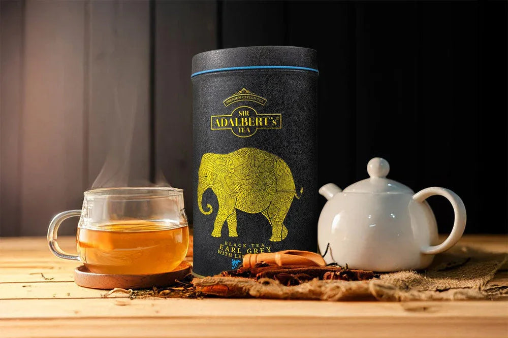 Herbata Earl Grey Sir Adalbert's w czarnej puszce z żółtym słoniem, szklanka parującej herbaty i biała czajnik ceramiczny