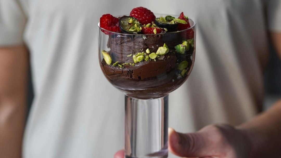 Mus czekoladowy w szklance ozdobiony malinami i posiekanymi pistacjami trzymany przez osobę w białej koszulce